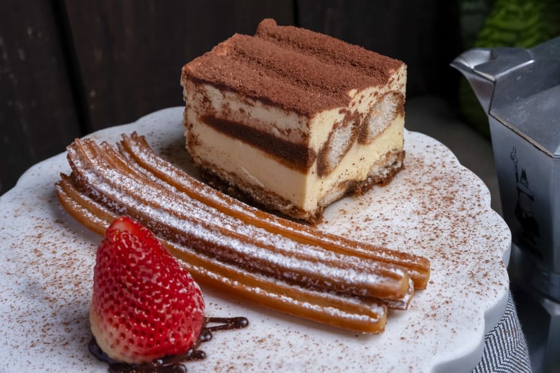 Tiramisu slice, powdered sugar churros, and a strawberry with chocolate drizzle on a white plate.