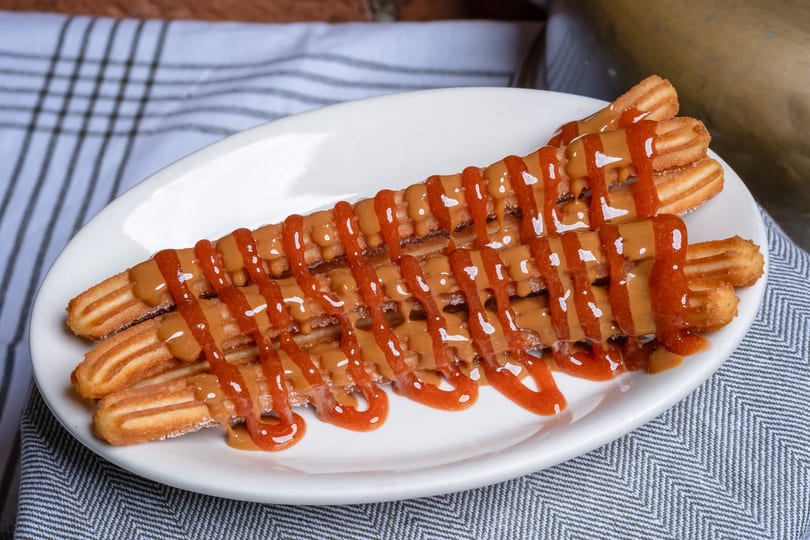 Golden churros on a white plate, drizzled with caramel and red fruit sauce on a patterned cloth.