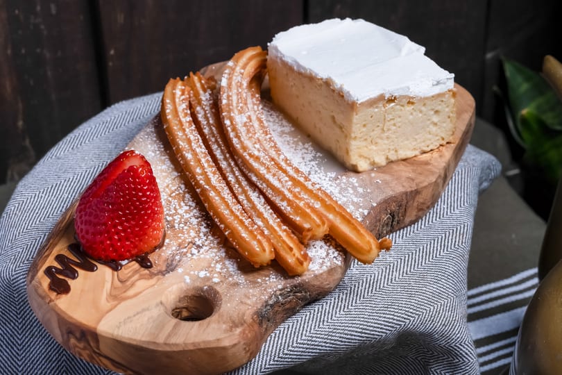 Churros, tres leches cake, and a strawberry with chocolate drizzle on a wooden board.
