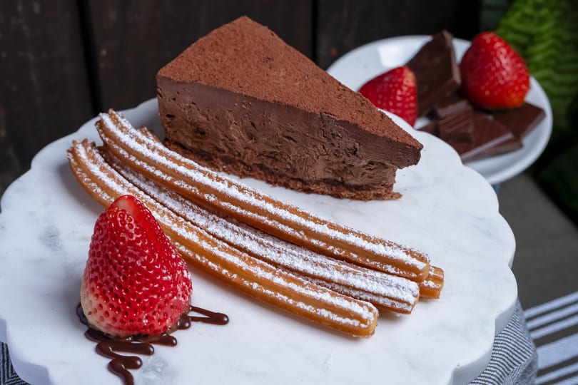 Decadent chocolate cake slice, powdered churros, and fresh strawberries on a white marble serving tray.