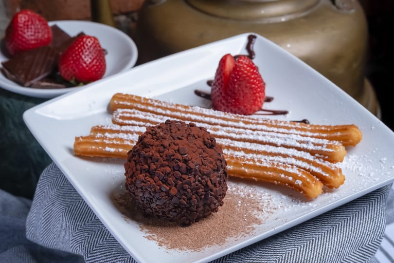 A dessert plate with powdered sugar churros, a chocolate truffle, and fresh strawberries.