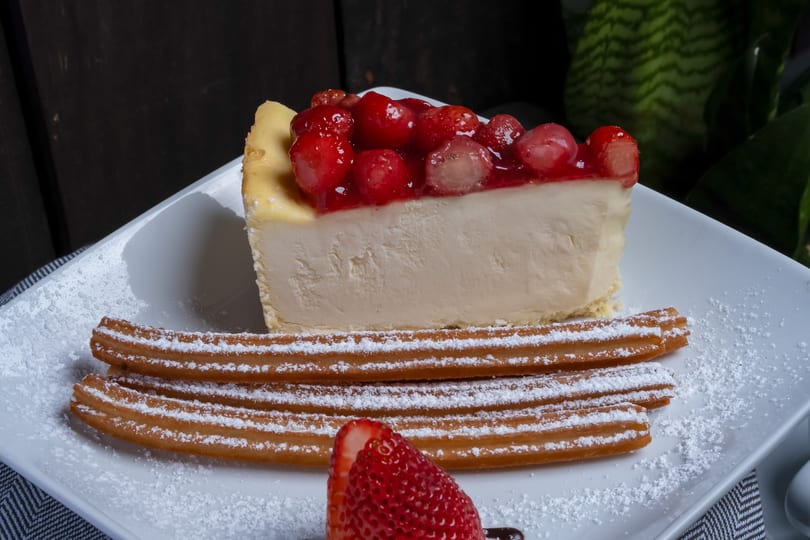 Strawberry cheesecake slice with churros and powdered sugar on a white plate.