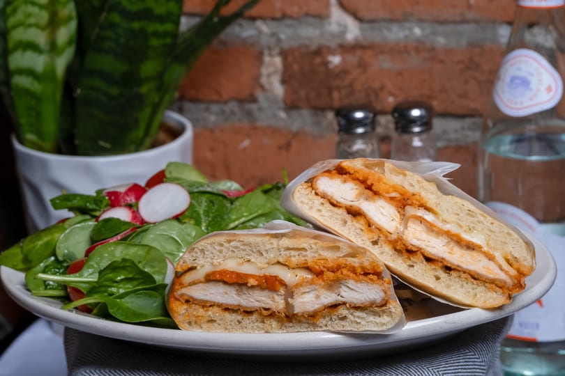 Chicken cutlet sandwich with melted cheese and sauce, served with a fresh spinach and radish salad.